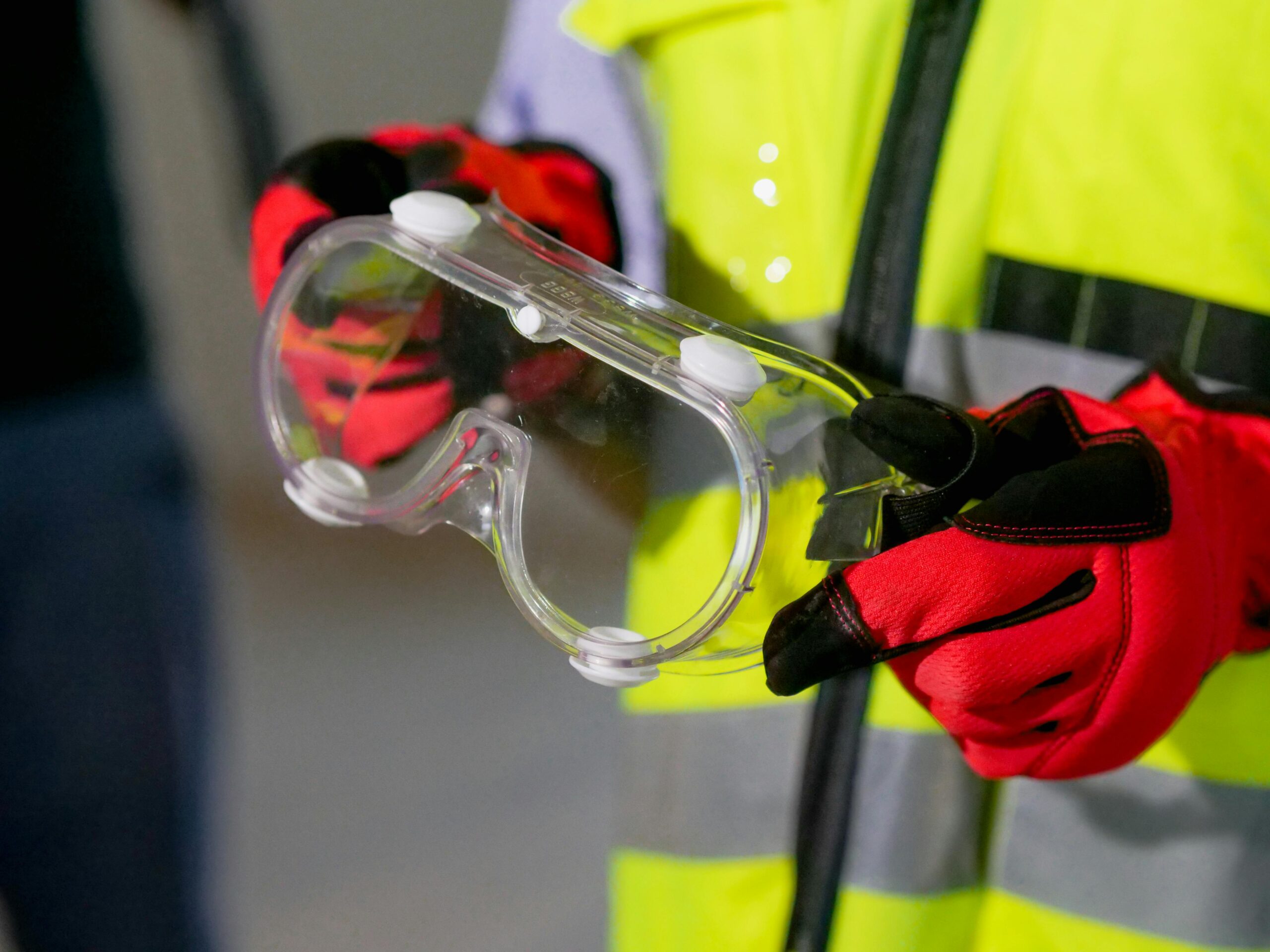 Close‑up van iemand die een veiligheidsbril vasthoudt met beschermende handschoenen, met zichtbare high‑visibility kleding.